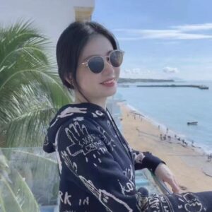 Relaxed young woman with short dark hair, wearing stylish sunglasses and a black hoodie with artistic white graphics, enjoying a sunny day by the ocean at Petal & Still.